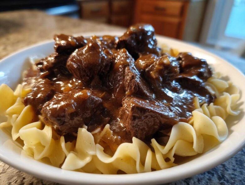 Close-up of tender crockpot beef tips smothered in rich brown gravy served over wide egg noodles in a white bowl.