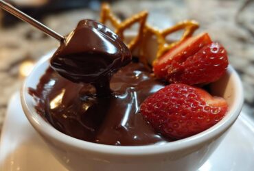 A piece of food being dipped into smooth, melted chocolate fondue, garnished with fresh strawberries and pretzels.