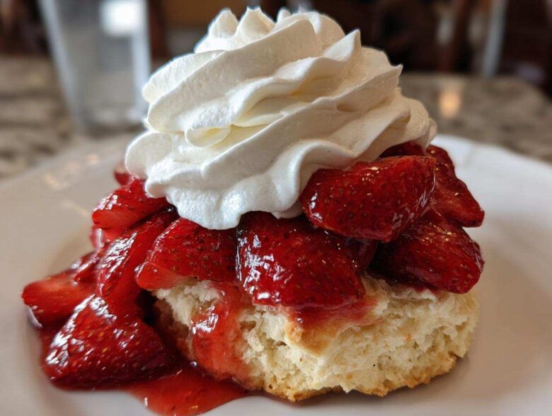 A close-up of a fluffy biscuit base topped with macerated strawberries and a generous swirl of whipped cream, perfect shortcake.