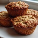 A close-up of four freshly baked, golden brown oatmeal muffins stacked slightly on a white plate.