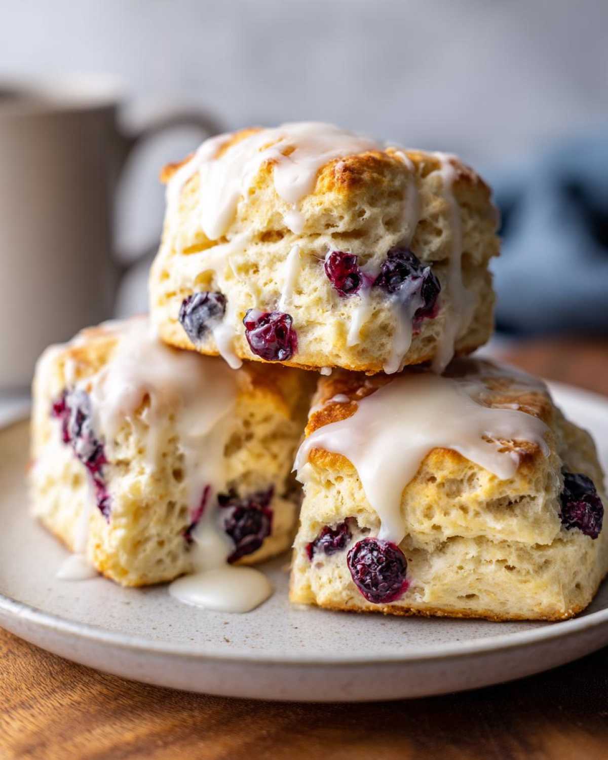 Three flaky blueberry scones stacked on a plate, generously topped with a dripping vanilla glaze.