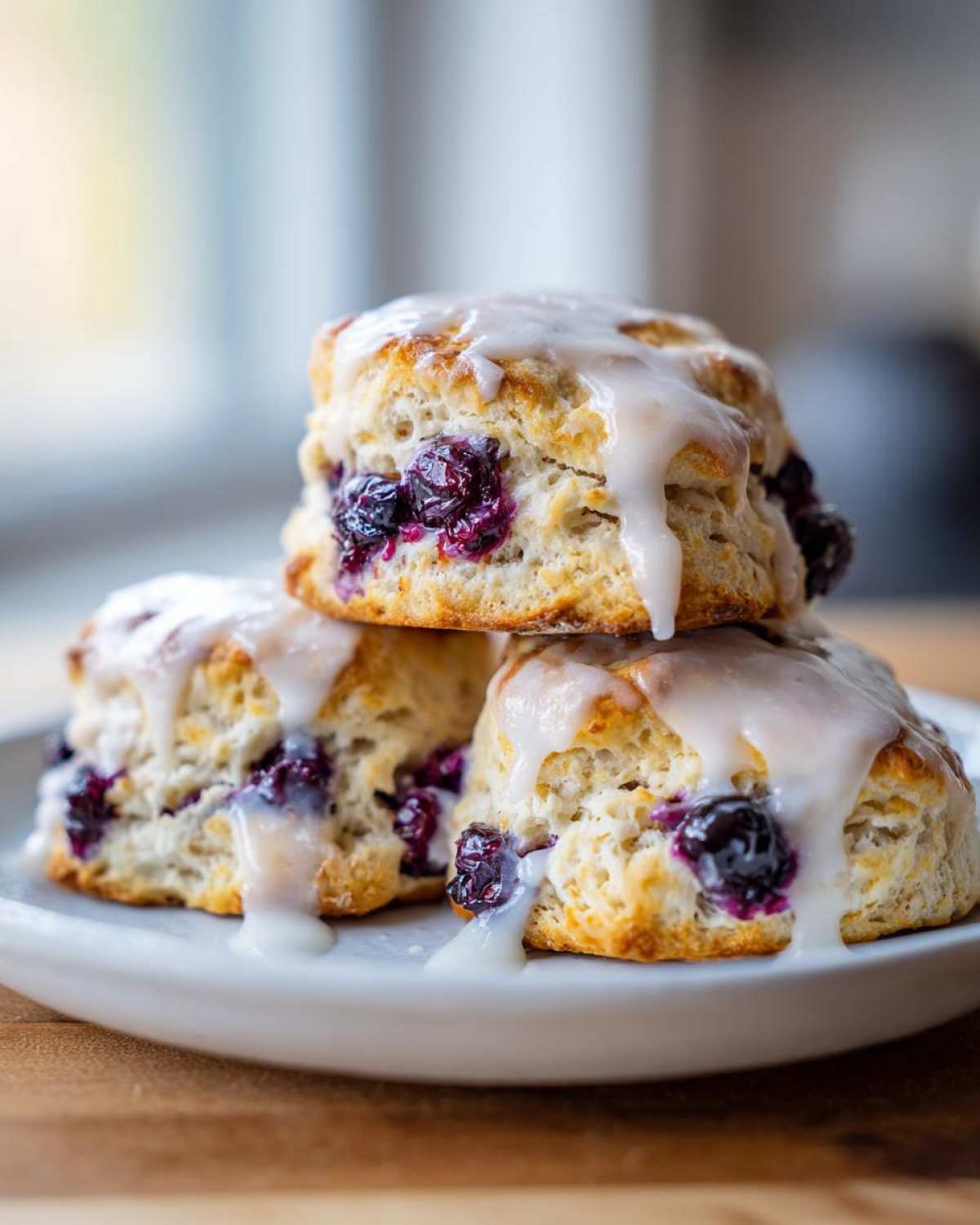 A stack of three flaky blueberry scones topped with a thick vanilla glaze, featuring visible bursts of dark blueberries.