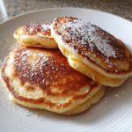 Three fluffy banana pancakes stacked on a white plate, generously dusted with powdered sugar.