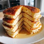 Close-up of a tall stack of fluffy pancakes with a slice removed, showing the airy interior and soaking in maple syrup.