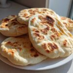 A stack of freshly cooked, fluffy pita bread with golden brown spots resting on a white plate.