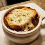 Close-up of a steaming bowl of french onion soup recipe topped with a melted Gruyère cheese crouton and cracked pepper.