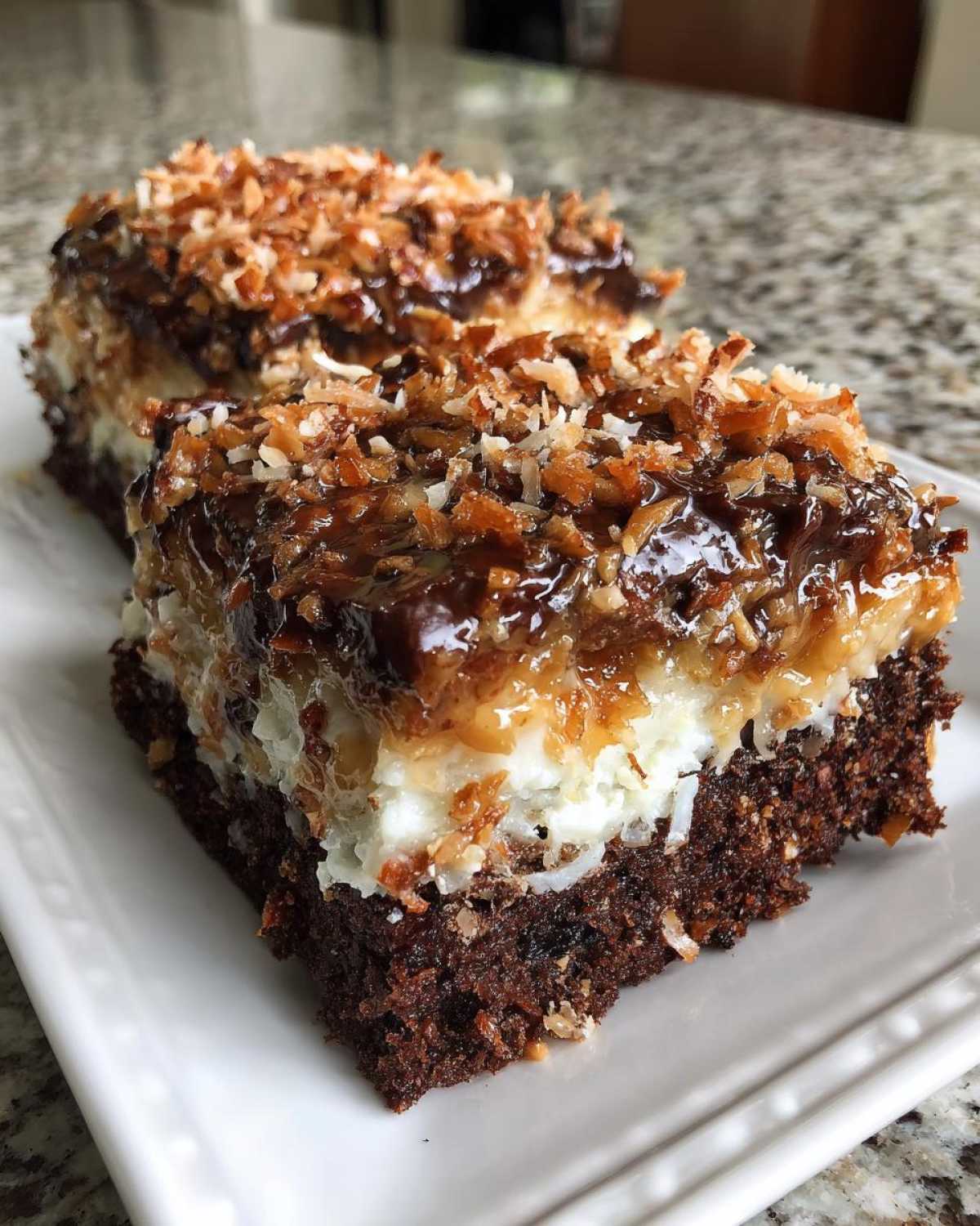 Close-up of a rich slice of german chocolate poke cake showing the chocolate base, white filling, and toasted coconut pecan topping.