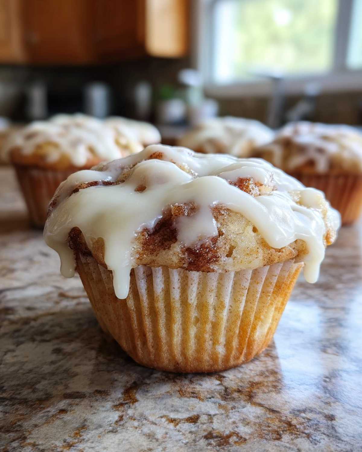 A close-up of one freshly baked cinnamon roll muffin topped with thick white icing, with more muffins blurred in the background.