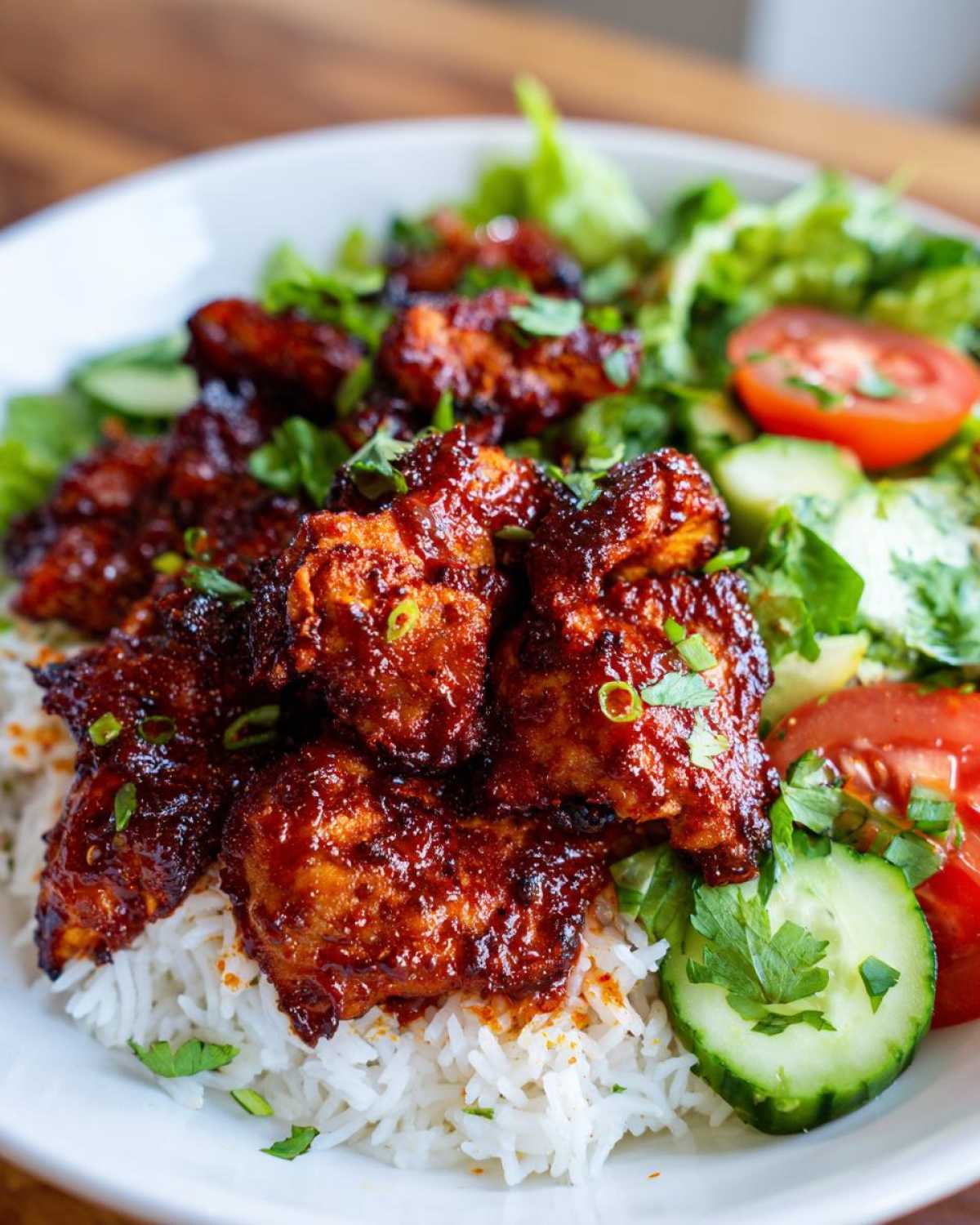 Close-up of a bowl featuring saucy, glazed harissa chicken served over white rice with fresh salad greens, cucumber, and tomato.