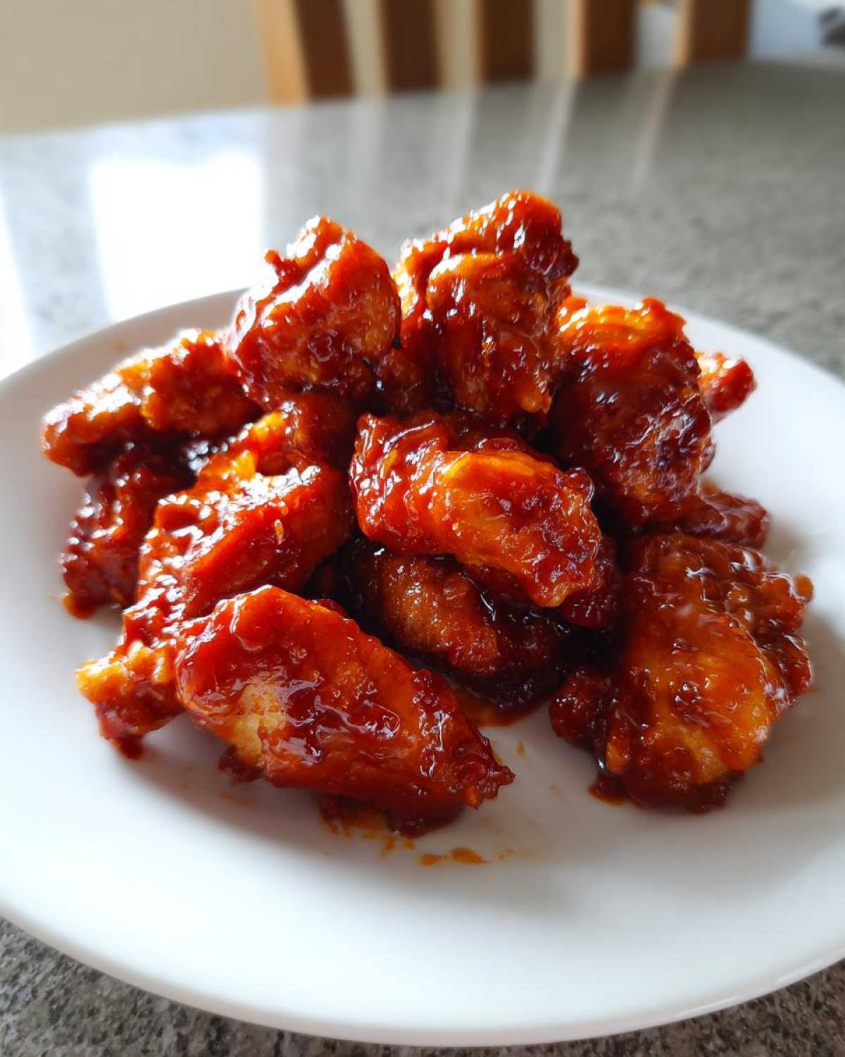 Close-up of crispy chicken pieces heavily coated in a shiny, deep red gochujang sauce served on a white plate.