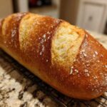 A golden-brown, freshly baked loaf of french bread with a crisp, scored crust resting on a black wire cooling rack.