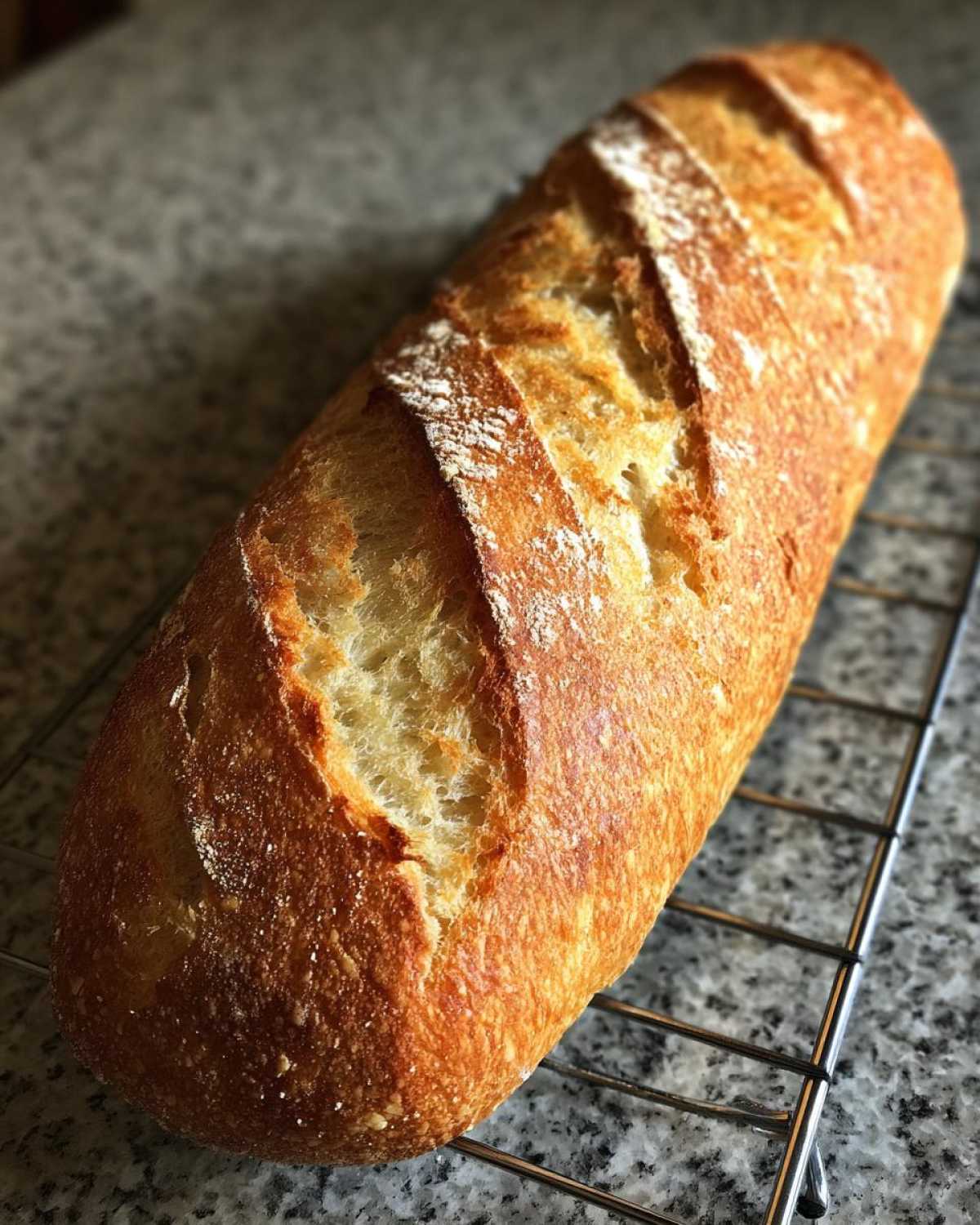 A freshly baked, golden-brown loaf of french bread with crisp slashes resting on a wire cooling rack.