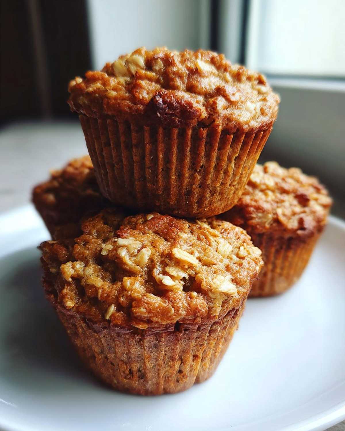 A stack of three freshly baked, golden brown oatmeal muffins with visible oat topping on a white plate.