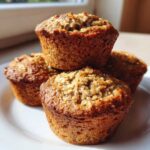 A stack of four freshly baked, golden brown oatmeal muffins with visible oat topping, sitting on a white plate.