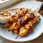 Close-up of grilled chicken satay skewers served on a white plate next to a small bowl of creamy peanut sauce.