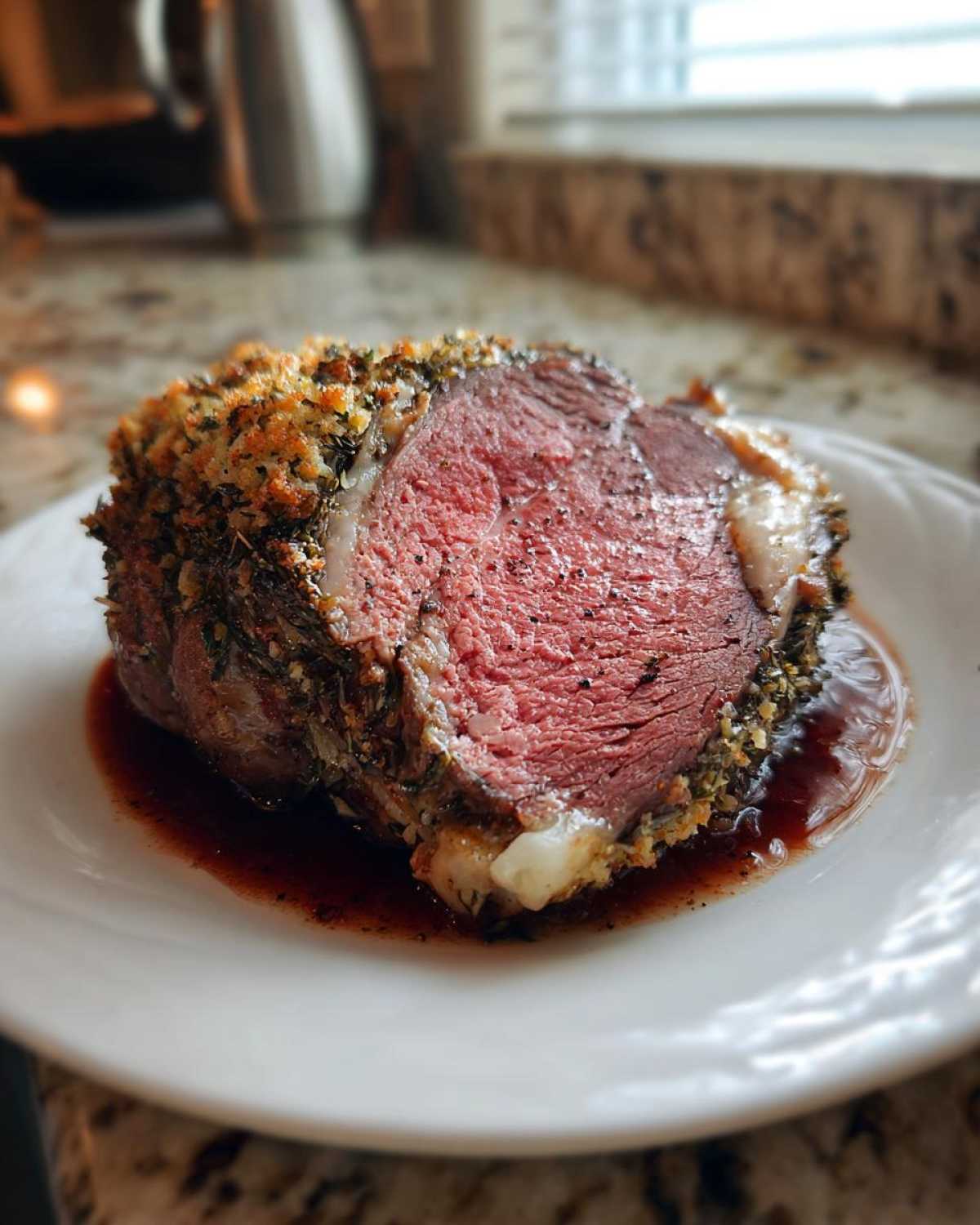 A thick slice of medium-rare prime rib recipe roast with a golden herb crust, resting in its juices on a white plate.