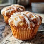 A close-up of a freshly baked cinnamon roll muffin topped with thick white icing drizzle.