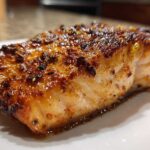 Close-up of a juicy, caramelized air fryer salmon fillet with a dark, seasoned crust resting on a white plate.