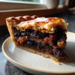 A close-up, sunlit slice of mincemeat pie showing flaky pastry and rich, dark fruit filling.