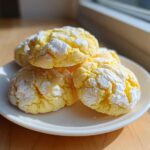 A stack of bright yellow lemon crinkle cookies dusted heavily with powdered sugar, sitting on a small white plate.