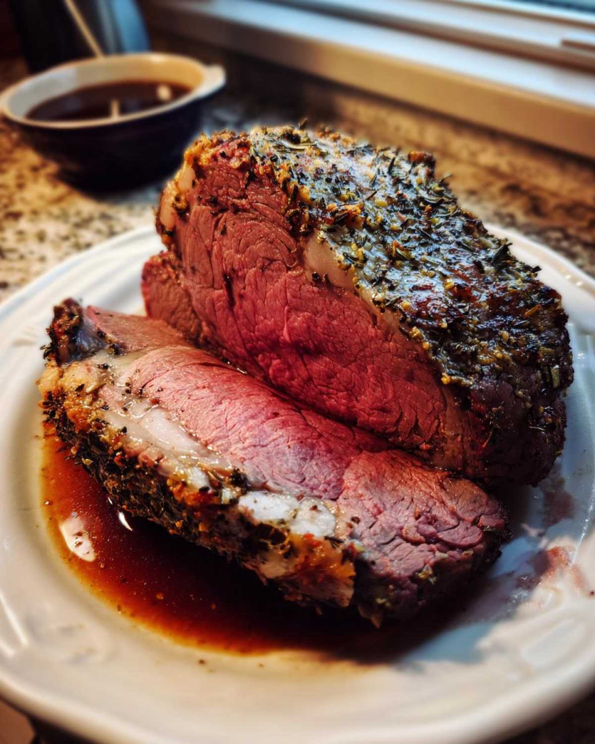 A thick slice of medium-rare prime rib recipe roast with a dark herb crust, resting in its juices on a white plate.