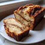 A moist apple bread loaf, sliced to show the cinnamon swirl and chunks of apple inside, resting on a white plate.