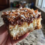 Close-up of a hand holding a moist slice of german chocolate poke cake topped with gooey caramel and toasted coconut.