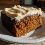 A close-up, sunlit slice of rich, dark gingerbread cake topped with thick, creamy frosting on a white plate.