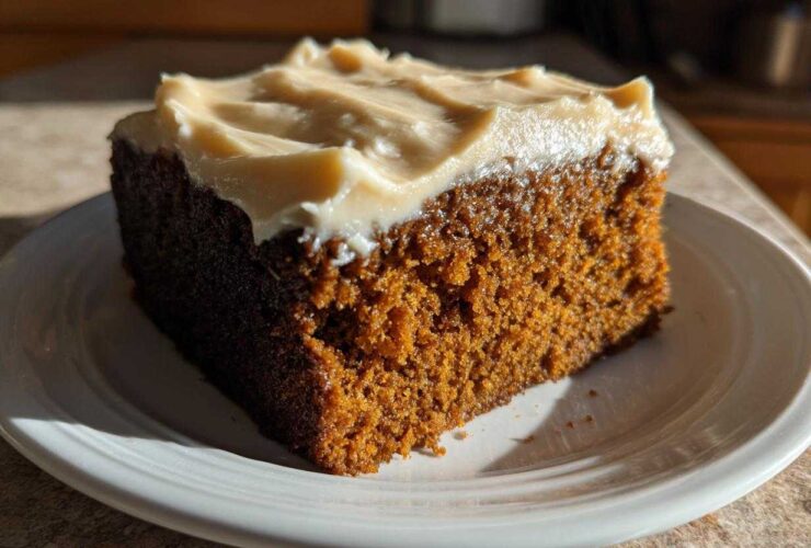 A close-up, sunlit slice of rich, dark gingerbread cake topped with thick, creamy frosting on a white plate.