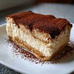 A rich slice of no bake tiramisu cheesecake layered with cream and dusted heavily with cocoa powder.