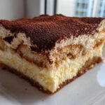 A perfect slice of no bake tiramisu cheesecake showing layers of cream, ladyfingers, and a cocoa powder topping.