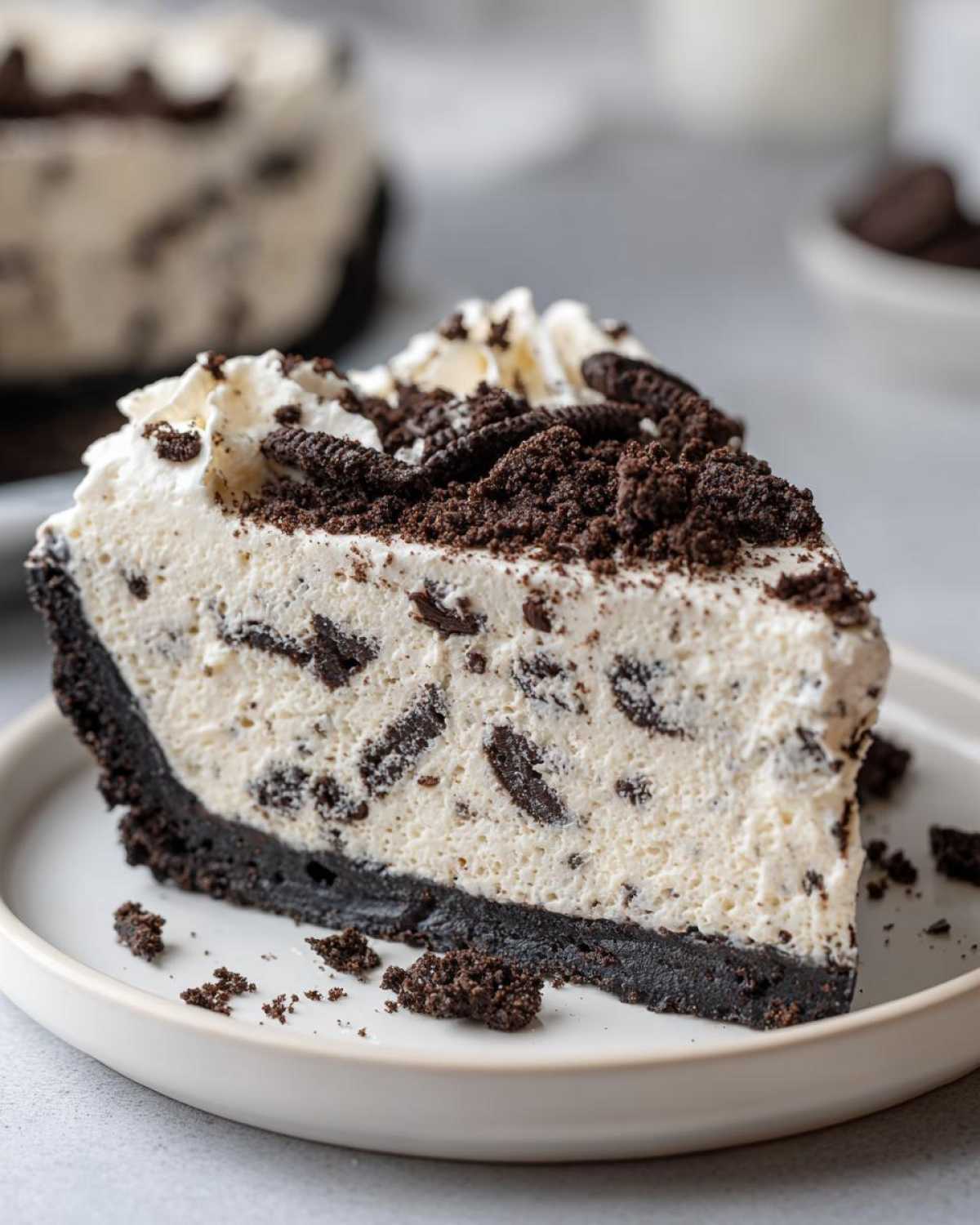 A close-up of a generous slice of no-bake Oreo pie featuring a dark crust, creamy filling studded with cookie pieces, and topped with whipped cream and crumbs.