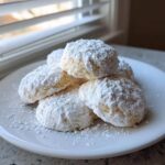 A stack of soft, powdered sugar-dusted Cool Whip Cookies piled high on a white plate near a window.