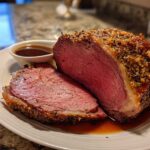 A thick slice of medium-rare prime rib recipe roast with a seasoned crust, resting on a plate with au jus.