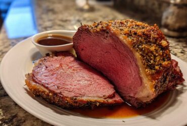 A thick slice of medium-rare prime rib recipe roast with a seasoned crust, resting on a plate with au jus.