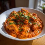 A close-up of rich, orange-red butter chicken pieces generously garnished with fresh cilantro.