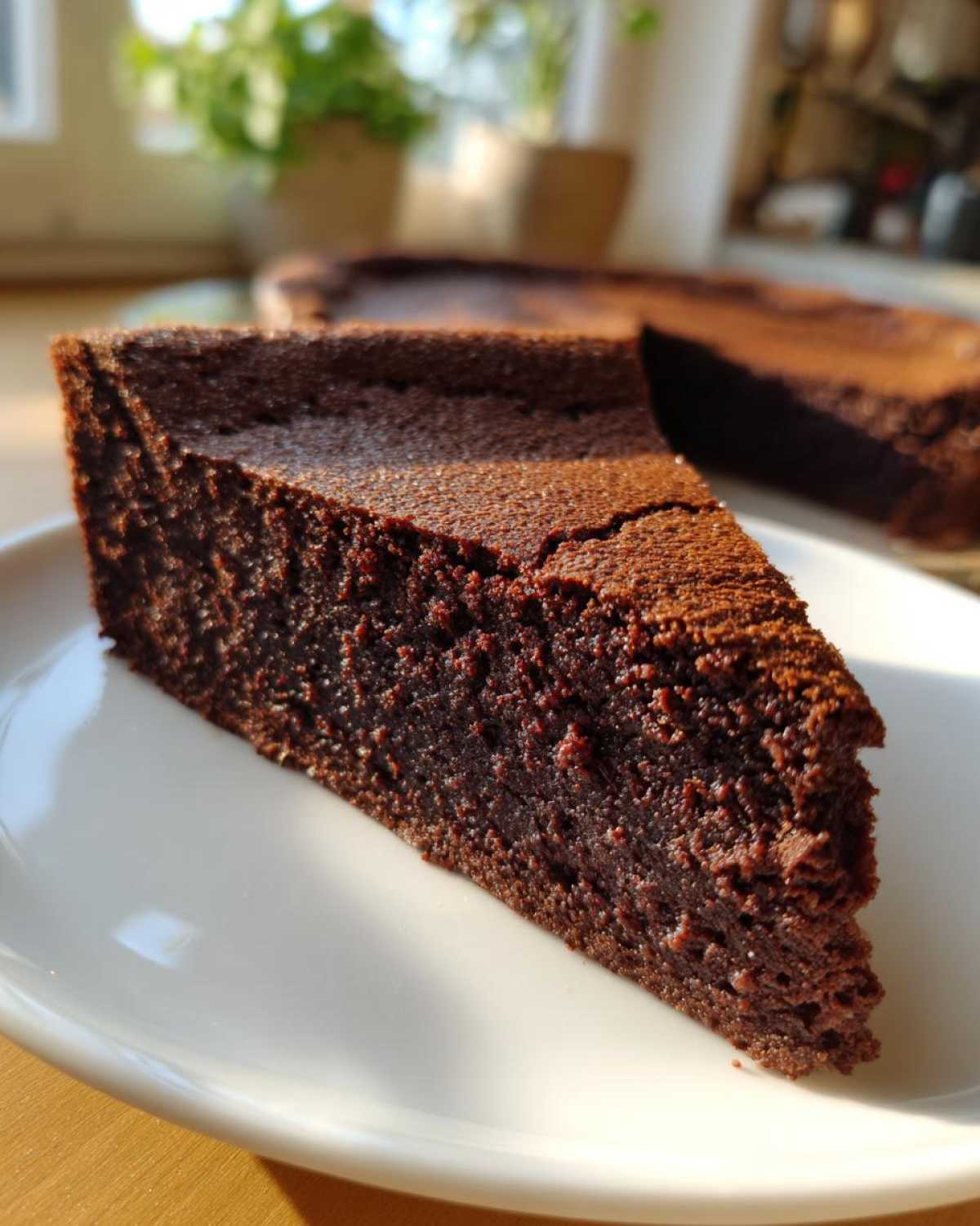 A close-up of a dense, dark slice of flourless chocolate cake resting on a white plate, with the rest of the cake blurred in the background.