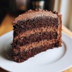 A close-up of a moist, multi-layered slice of chocolate cake recipe with thick chocolate frosting.
