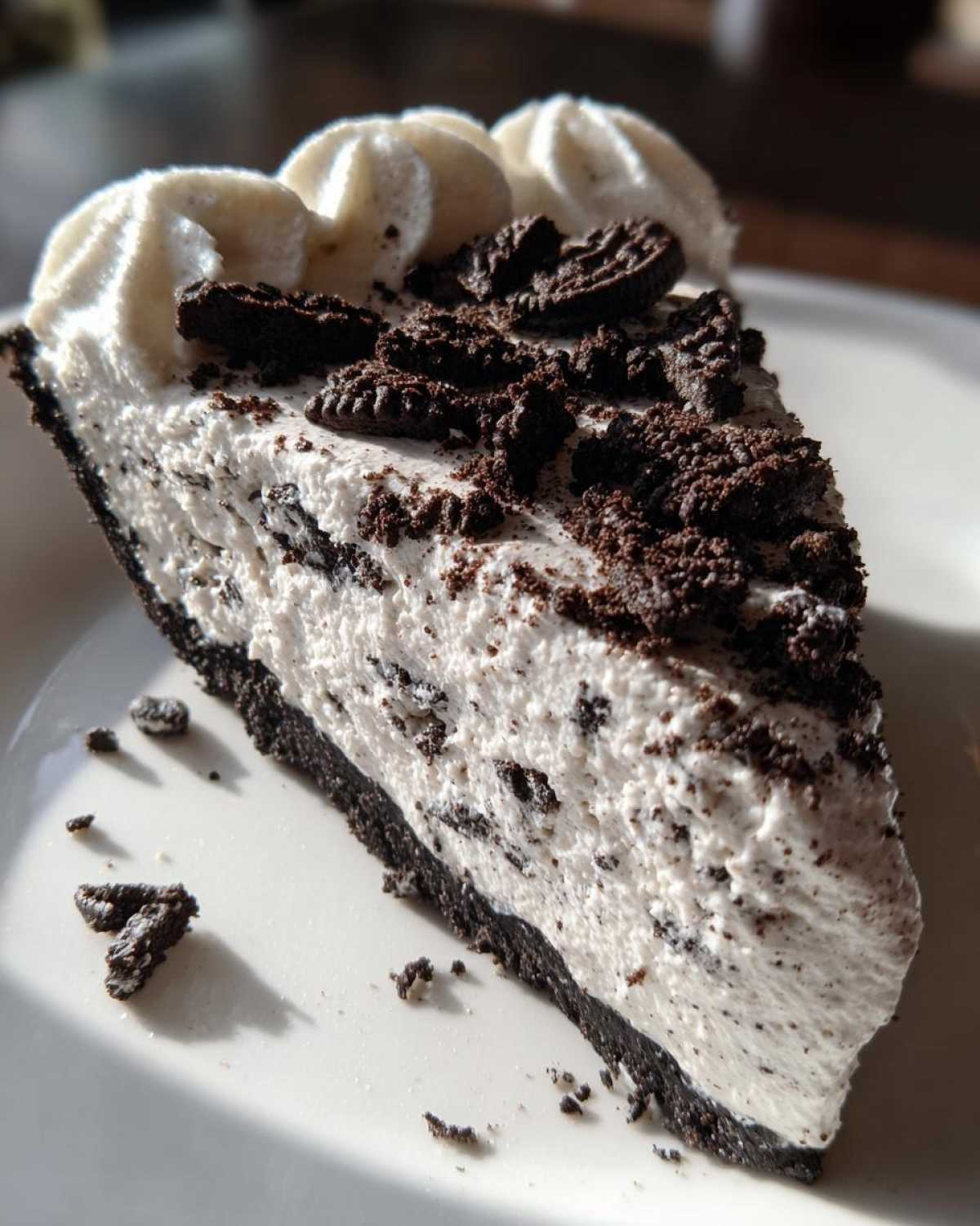 A close-up slice of rich, creamy Oreo pie with a dark chocolate crust, topped with whipped cream and cookie crumbles.