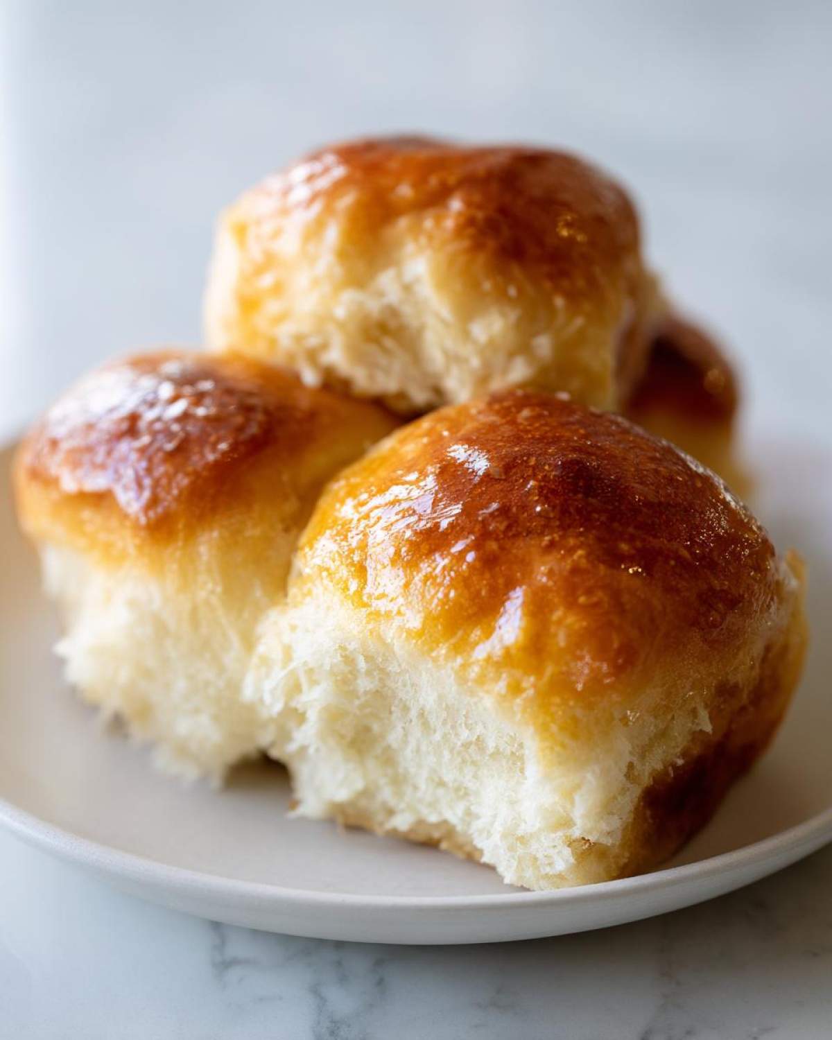 Close-up of three fluffy sourdough discard rolls stacked on a white plate, showing a shiny, golden-brown butter glaze.