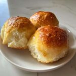 Three fluffy, golden-brown sourdough discard rolls with a shiny glaze resting on a small white plate.