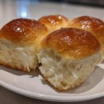 Four golden brown, fluffy sourdough discard rolls with a shiny glaze resting on a white plate.