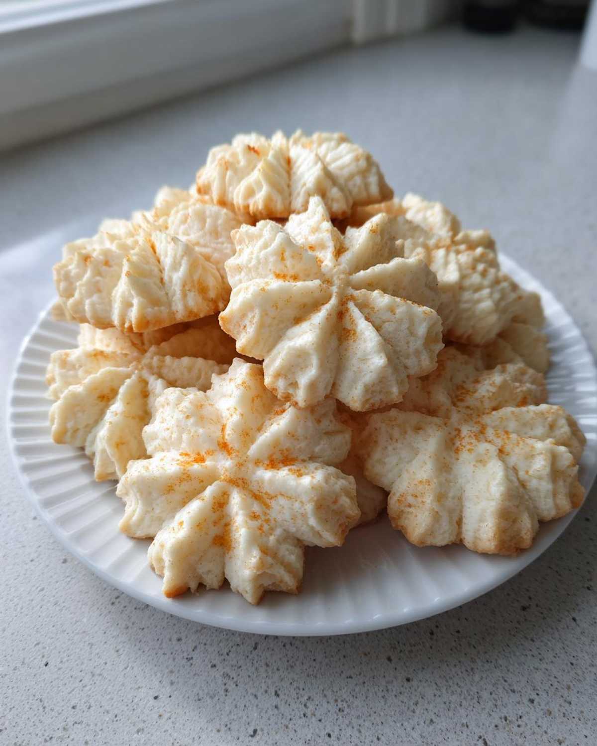 A mound of delicate, star-shaped whipped shortbread cookies lightly dusted with orange spice on a white plate.