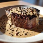 A thick, seared steak generously covered in creamy peppercorn sauce and whole peppercorns.