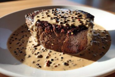 A thick, seared steak generously covered in creamy peppercorn sauce and whole peppercorns.