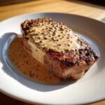 A perfectly seared steak generously topped with creamy peppercorn sauce and whole peppercorns.