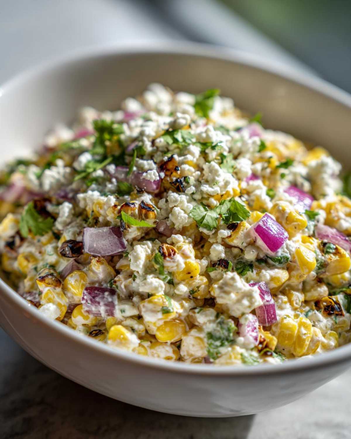 A close-up view of a creamy street corn salad featuring charred corn, red onion, cotija cheese, and cilantro.