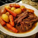 A close-up of tender, shredded pot roast served with roasted carrots and small yellow potatoes in rich gravy.