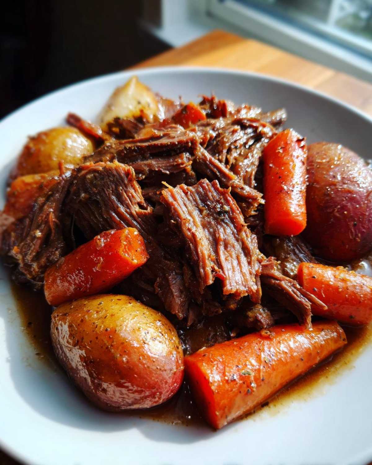 Close-up of tender, shredded pot roast served with large chunks of carrots and small potatoes in rich gravy.