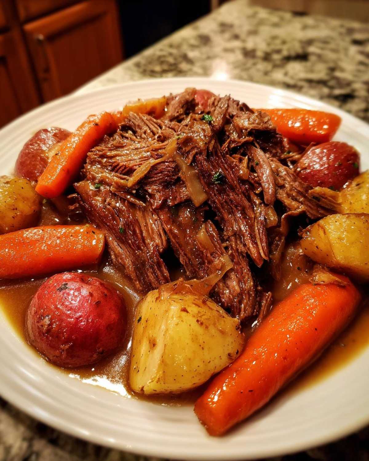 A close-up of a plate featuring tender, shredded pot roast surrounded by cooked carrots and red potatoes in rich gravy.